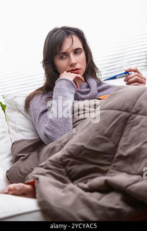 Die kranke Frau liegt im Bett, in eine Decke gewickelt, überprüft ihre Temperatur mit einem Thermometer, kämpft zu Hause gegen Krankheiten Stockfoto