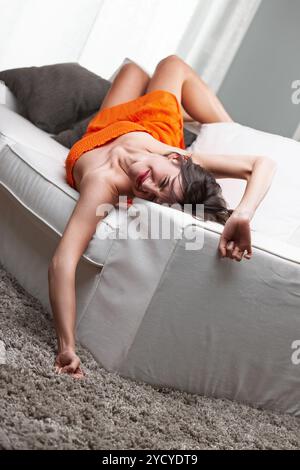 Die junge Frau streckt sich auf einem weißen Sofa, ihr Arm hängt zum Boden hin. Sie scheint entspannt zu sein, ihr Ausdruck zeigt leichte Zufriedenheit, Sugar Stockfoto