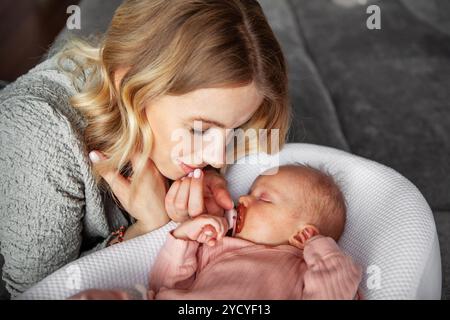 Die schöne Mutter blickt mit Liebe auf das schlafende Neugeborene, das in rosa Einteiler gekleidet ist und einen Schnuller im Mund hat. Ein ruhiger Moment zu Hause, spiegelt tiefe Verbundenheit wider Stockfoto