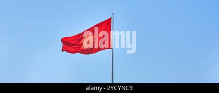 Banner Kirgisistan aktualisierte Flagge flattert gegen den klaren blauen Himmel. Rote neue Flagge der Kirgisischen Republik auf dem Fahnenmast schwenken, echtes Foto. Kirghizia Stockfoto