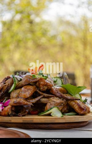 BBQ Hähnchenflügel garniert mit Mikrogrün, Blumen und frischen Gurkenscheiben. Wunderschönes Servieren auf einem Holzteller. Seitenansicht. Ästhetisches Abendessen im Freien Stockfoto