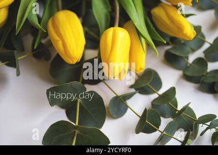 Schöne gelbe Tulpen mit Eukalyptuszweigen auf einem weißen Tisch, Inschrift Happy day Stockfoto