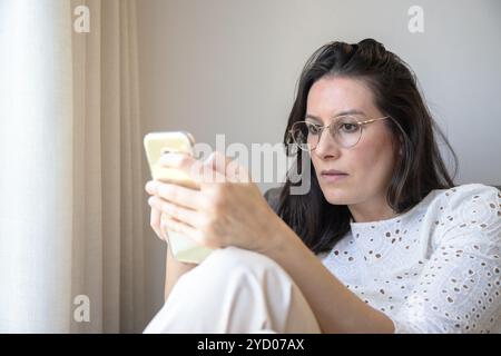 Eine Frau mit Brille schaut konzentriert auf ihr Smartphone. Drinnen neben einem Vorhang zu sitzen, scheint sie in den Bildschirm eingetaucht zu sein. Stockfoto