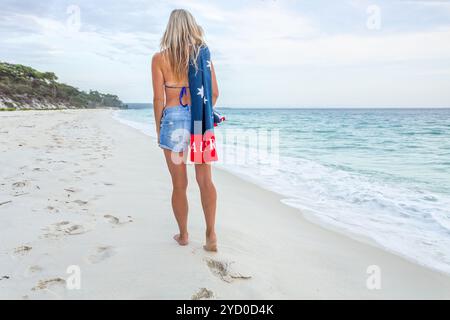 Am frühen Morgen läuft eine Frau barfuß am Strand entlang. Sie trägt Denim-Shorts, Bikini und ein Handtuch über der Schulter Stockfoto