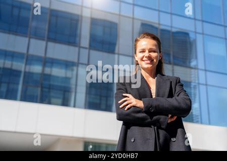 Geschäftsfrau im schwarzen Mantel steht mit überkreuzten Armen und lächelt vor einem Bürogebäude. Stockfoto