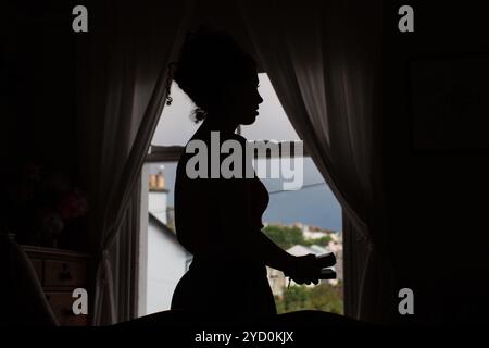 Silhouette einer Frau an einem Fenster mit Vorhängen, das ein Objekt hält, vor einem weichen, natürlichen Hintergrund und einem bewölkten Himmel. Stockfoto