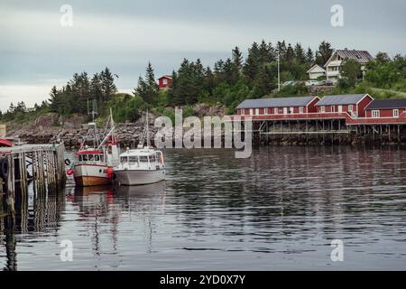 Zwei Fischerboote ruhen friedlich an einem hölzernen Dock an einem ruhigen Abend auf den Lofoten-Inseln, umgeben von üppigem Grün und roten Gebäuden am Ufer. Stockfoto