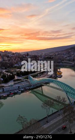 Die Friedensbrücke, die bei Sonnenuntergang in Tiflis, Georgien, beleuchtet wird, überspannt den Fluss Kura mit dramatischem orange-rosa Himmel über der Hauptstadt. Stockfoto