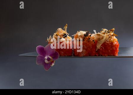 Elegante Sushi-Brötchen, serviert auf einer Messerklinge mit Orchideenblume vor dunklem Hintergrund Stockfoto