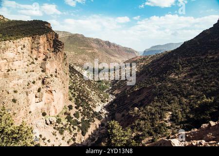 Ein atemberaubender Blick auf die marokkanischen Badlands zeigt steile Abhänge und eine gewundene Schotterstraße durch das Tal, flankiert von Kaparral und verstreut Stockfoto