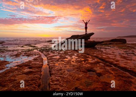 Frau, die auf Küstenfelsen steht, mit atemberaubendem Sonnenaufgang und Reflexionen Stockfoto
