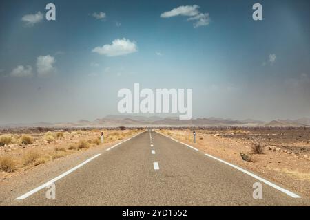 Eine lange, leere Asphaltstraße führt durch die trockene, beige Landschaft der marokkanischen Wüste. Cumulus-Wolken ziehen über sich und verstärken die warme, sonnige Atmosphäre Stockfoto