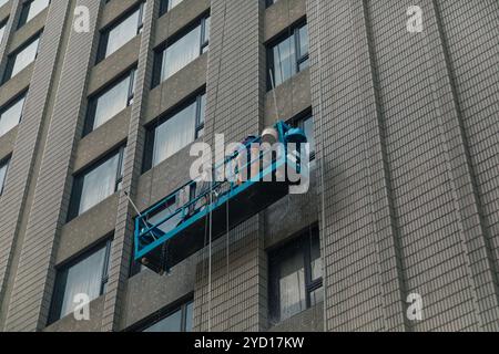 Zwei Arbeiter werden auf einem Bahnsteiglift aufgehängt, um die Fenster eines hohen Gebäudes in Taipeh zu reinigen. Die Umgebung zeigt moderne Architektur unter hellem Himmel Stockfoto