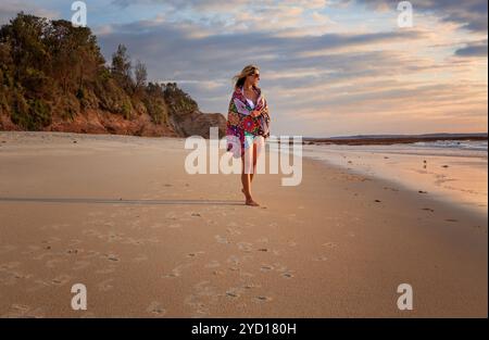 Eine Frau im Bikini mit einem Strandtuch, das locker um sie gewickelt ist, läuft am frühen Morgen am Strand entlang, mit einer sanften Brise im Haar, während sie zusieht Stockfoto