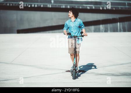 Ein junger Mann mit lockigen Haaren steht auf einem Elektroroller, trägt ein hellblaues Denim-Hemd und khakifarbene Shorts. Er schaut gegen einen Minimalis zur Seite Stockfoto
