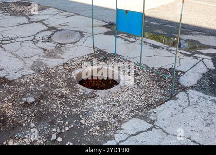 Ungesicherte Abwasserkanal Mannloch auf der Asphaltstraße öffnen. Gefährliche Grube auf der Straße. Alte Kanalisation gut Klappe auf der Straße Stockfoto