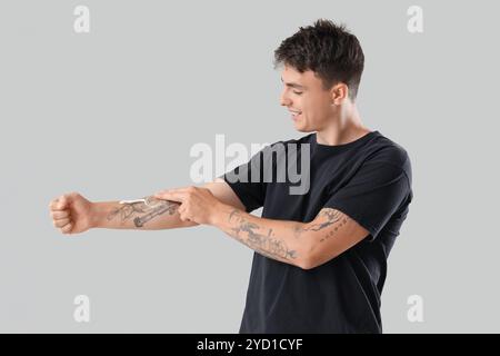 Junger Mann, der Creme auf seinem tätowierten Arm vor hellem Hintergrund aufträgt Stockfoto
