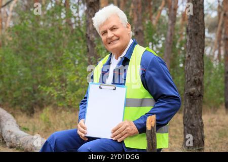 Leitender Forstwirt mit leerem Klemmbrett, der auf Baumstamm im Wald sitzt Stockfoto