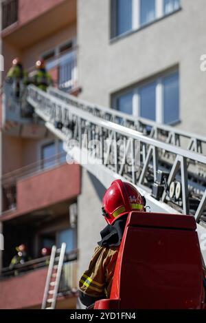 Der Bediener eines Feuerwehrwagens mit verlängerter Hochleiter reicht bis zum Wohngebäude, in dem Feuerwehrleute in Schutzausrüstung auf dem Balkon arbeiten Stockfoto