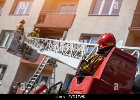 Der Bediener eines Feuerwehrwagens mit verlängerter Hochleiter reicht bis zum Wohngebäude, in dem Feuerwehrleute in Schutzausrüstung auf dem Balkon arbeiten Stockfoto