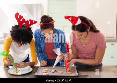 Multirassische Freundinnen, die weihnachtskekse schmücken, festliche Stirnbänder tragen, zu Hause Stockfoto