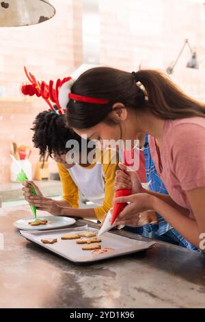 Festliche Stirnbänder tragen, multirassische Freundinnen, die weihnachtskekse schmücken, zu Hause Stockfoto