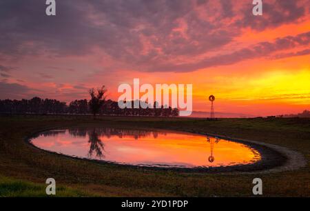 Windmühle an einem Teich in ländlichem Feld mit leuchtendem roten, orangen und gelben Sonnenaufgang und einem leichten, tief liegenden Nebel. Ländliches Australien Stockfoto