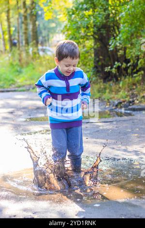 Ein kleiner Junge springt in eine Pfütze. Ein Junge in Gummistiefeln. Glückliche Kindheit. Pfützen nach dem Regen. Warmer Sommerabend. Spiele für Kinder Stockfoto