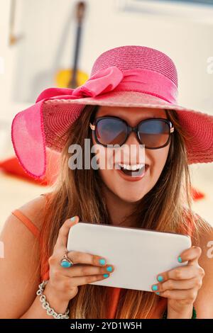 Eine junge Frau mit rosafarbenem Sonnenhut und Sonnenbrille genießt ihr digitales Tablet zu Hause, taucht in Unterhaltung ein und bleibt drahtlos in Verbindung Stockfoto