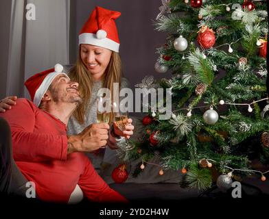 Ein Paar feiert den Weihnachtsanfang, neben dem Baum, mit 2 Gläsern Champagner, Weihnachtsmützen, lächelnd zu Hause Stockfoto