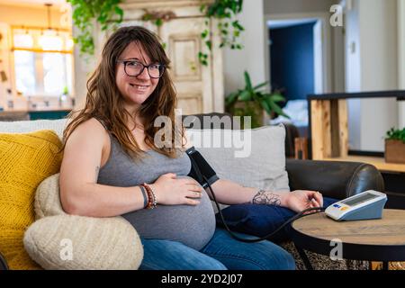 Fröhliches Porträt einer jungen Frau in späteren Schwangerschaftsstadien, die routinemäßige Gesundheitskontrollen zu Hause durchführt und ein Blutdruckmessgerät mit Armmanschette trägt Stockfoto