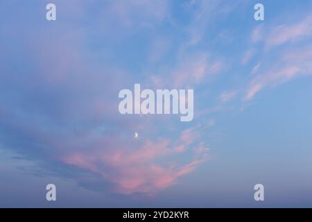 Pastellfarbener westlicher Himmel mit Halbmond. Ein ruhiger, pastellfarbener Himmel bei Sonnenuntergang mit sanften rosa Wolken, die über einem blauen Hintergrund schweben, und einem schwachen Mondsichel V Stockfoto