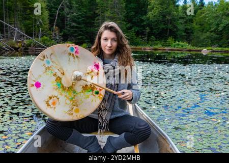 Schöne junge schamanische Frau, die im Kanu sitzt und eine heilige einheimische Rahmentrommel mit einem mit Pelz überzogenen Stock im See von Kanada mit Wasserlilien spielt Stockfoto