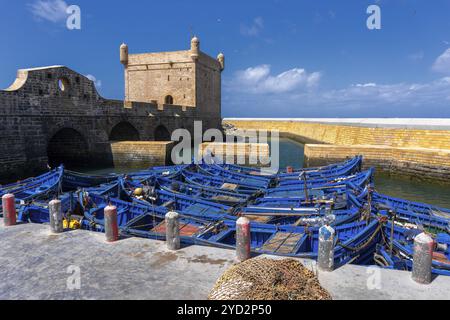 Essaouira, Marokko, 26. März 2024: Der Wachturm Borj el Barmil und farbenfrohe blaue Fischerboote im alten Hafen von Essaouira, Afrika Stockfoto