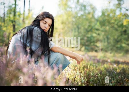 Eine junge Frau sitzt ruhig auf dem Waldboden, ihr langes Haar zieht sich über den Rücken. Sie erscheint tief in Gedanken, umgeben von blühenden Wildblumen Stockfoto