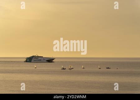 Ciutadella, Spanien, 26. Januar 2024: Kleine Segelboote vor einer Balearenfähre, die bei Sonnenuntergang in den Hafen von Ciutadella einfahren Stockfoto