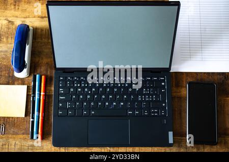 Ein Laptop wird mit einem Stift, einem Mobiltelefon und einem Hefter auf einem Schreibtisch abgelegt. Der Laptop ist geöffnet und der Bildschirm ist leer. Der Schreibtisch ist überfüllt mit verschiedenen Stockfoto