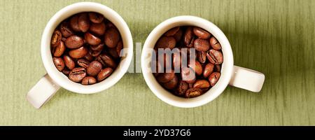 Zwei weiße Tassen mit gerösteten Kaffeebohnen werden auf einem grünen Hintergrund platziert. Die Lagerringe sind nebeneinander angeordnet, eine symmetrische Zusammensetzung. Stockfoto