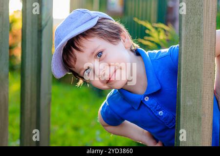 Der Junge schaut hinter dem Zaun raus. Der Junge trifft die Gäste. Lächelndes Kind. Wunderschönes Porträt eines Jungen. Artikel über ch Stockfoto