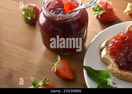 Glas mit Erdbeermarmelade auf Holztisch voller Früchte und weißer Teller mit Brotscheibe mit Marmelade. Erhöhte Aussicht. Stockfoto