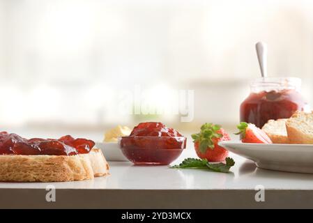 Frühstück mit Erdbeermarmelade mit Brotscheiben auf einer weißen Küchenbank und Küche im Hintergrund. Vorderansicht. Stockfoto