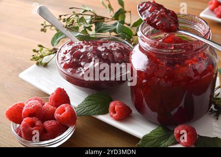 Detail der Gläser gefüllt mit Himbeermarmelade auf einem weißen Teller mit Blättern auf einem Holztisch mit einer Schüssel voller Beeren. Von oben erhobene Aussicht. Stockfoto