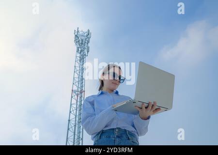 Frau, die an einem klaren Tag an einem Laptop in der Nähe eines Telekommunikationsturms arbeitet und Technologie und Konnektivität zeigt. Stockfoto