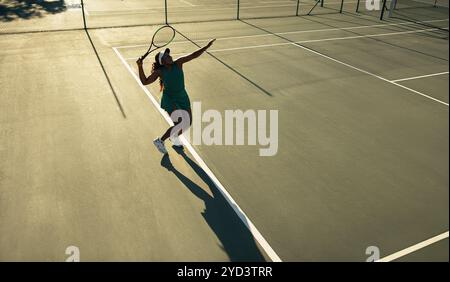 Eine Tennisspielerin, die bei Sonnenuntergang auf einem Platz im Freien eine Rolle spielt, wird als hintergrundbeleuchtete Silhouette erfasst. Stockfoto