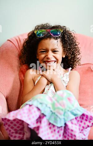 Lächelndes Mädchen mit bunter Sonnenbrille auf einem pinkfarbenen Stuhl, j Stockfoto