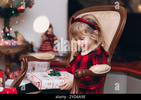 Ein kleines Mädchen im roten Weihnachtsoutfit öffnet zu Hause ein Geschenk Stockfoto