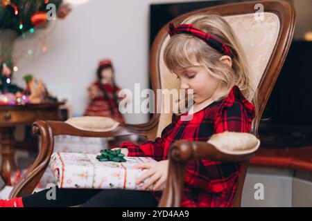 Ein kleines Mädchen im roten Weihnachtsoutfit öffnet zu Hause ein Geschenk Stockfoto