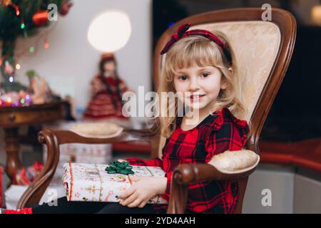 Ein kleines Mädchen im roten Weihnachtsoutfit öffnet zu Hause ein Geschenk Stockfoto