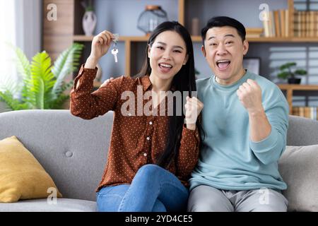 Ein aufgeregtes asiatisches Paar feiert den neuen Hauskauf und zeigt die Schlüssel, während er auf dem Sofa sitzt. Fröhliche Ausdrucksformen und Leistungsgefühl spiegeln Glück, Erfolg und gemeinsame Neuanfänge wider. Stockfoto