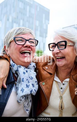 Zwei lachende, nur Erwachsene, kaukasische Freundinnen, die glücklich zusammen in der Stadt stehen. Stockfoto